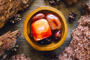 A collection of polished red gemstones sit elegantly in a wooden bowl, surrounded by rustic bark and natural elements, creating a warm, earthy ambiance.