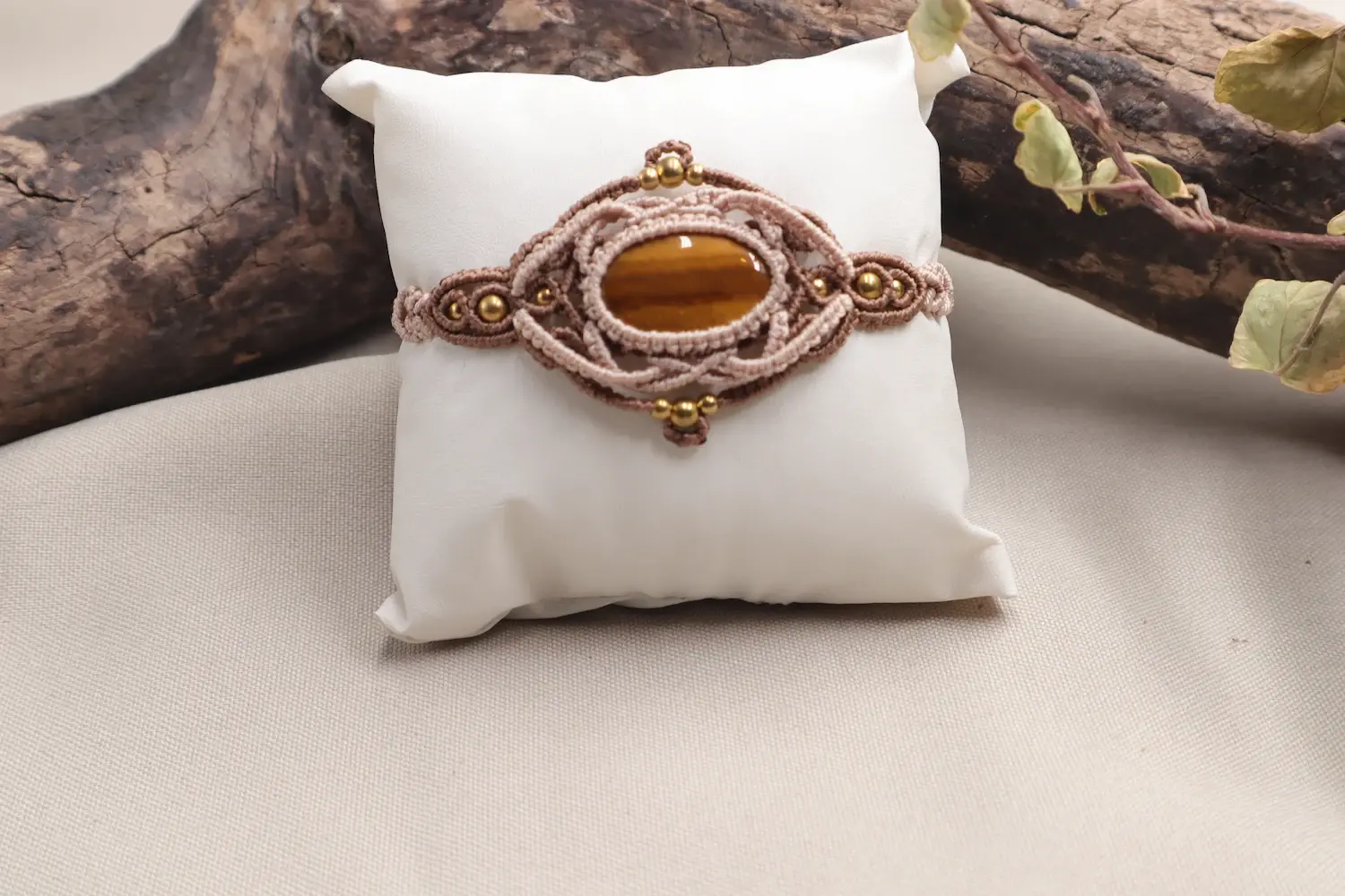 Bracelet en macramé marron et beige avec œil de tigre, modèle Mémoire de chêne, présenté sur coussin devant un bois flotté