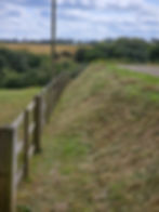 A grassy slope with a wooden fence and a field beyond the fence