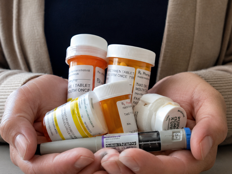 A woman’s hands cradle several prescription pill bottles and a used injection pen over a plain kitchen table in soft daylight, with her face out of frame.