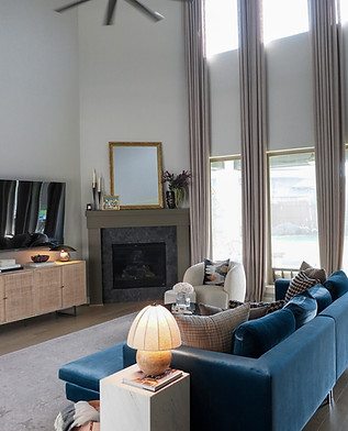 A modern living room with tall windows and floor-length curtains features a blue velvet sofa, a cream armchair, and a black f