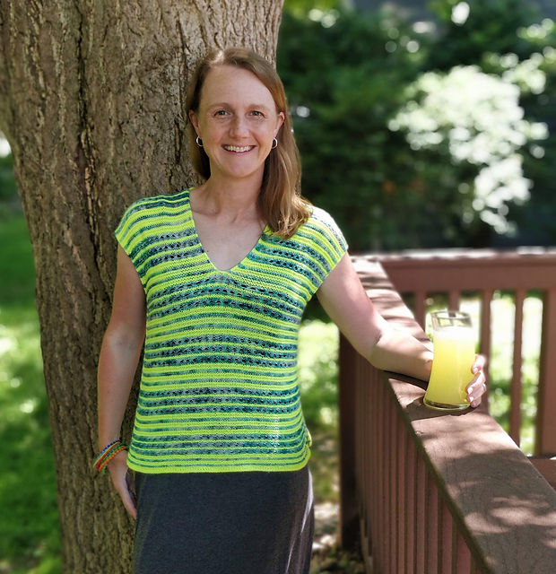 A woman stands wearing a yellow and blue v-neck, hand-knit, cap-sleeved top with horizontal stripes of alternating width.