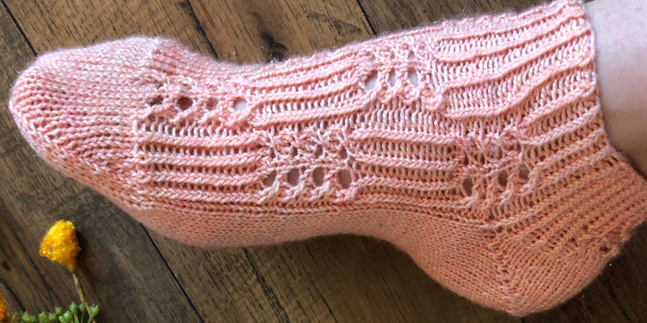 Pink knitted sock with lace patterns on a foot, resting on a wooden floor. Yellow flowers partially visible in the corner.
