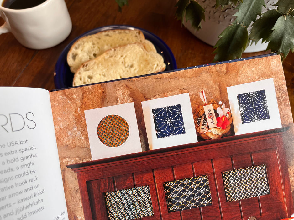 Open book showing intricate patterns on a wooden cabinet. Background has bread slices on a blue plate and a cup of coffee on a table.