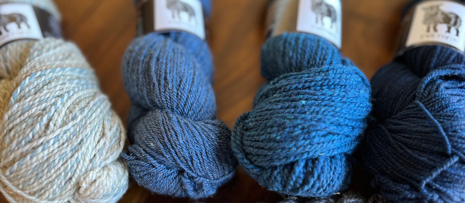 Five skeins of yarn in blue and gray tones are arranged on a wooden surface. Each skein has a label featuring an animal graphic.