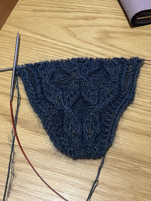 Blue lace knitting project with intricate pattern on wooden table. Circular knitting needle with red cable attached to the project.