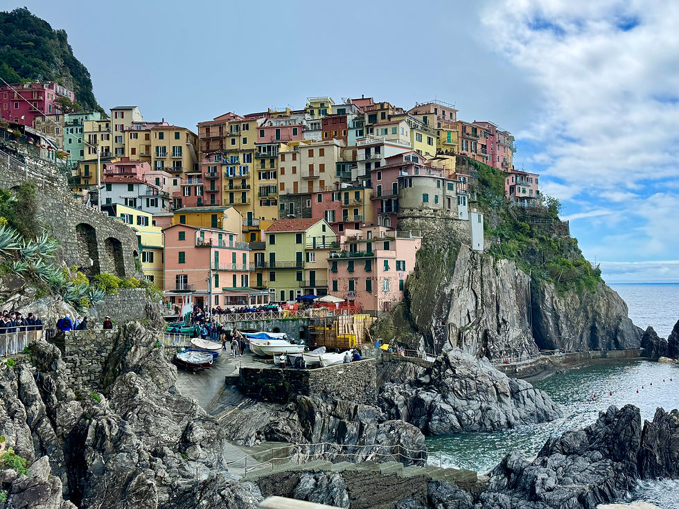 Colorful hillside village with pastel buildings on rocky cliffs by the sea. People walk along the stone path; boats are docked nearby.