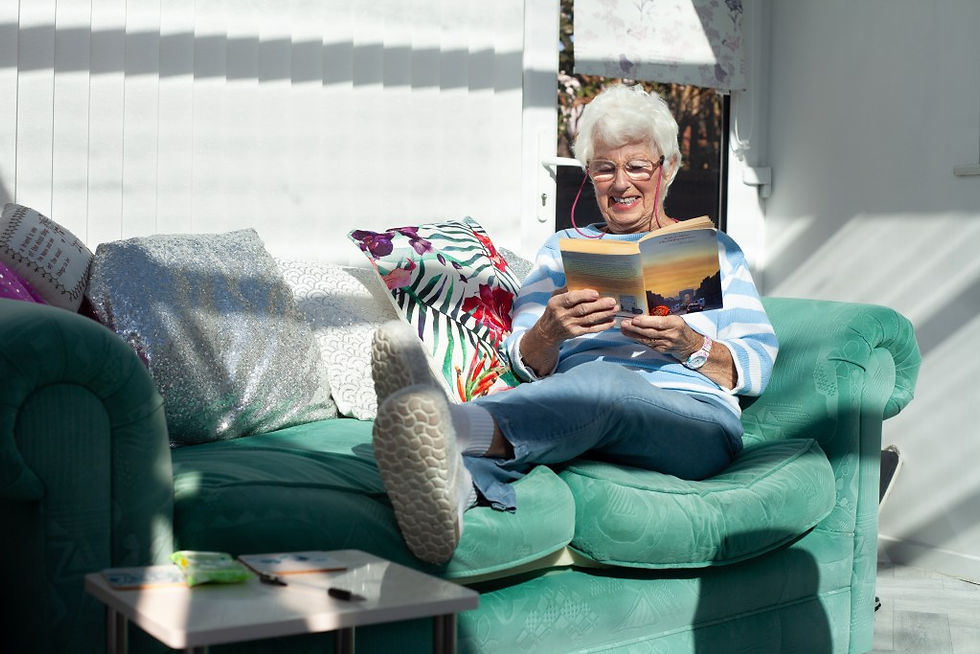 Elderly woman smiling, sitting on a green sofa reading a book. Bright sunlight, colorful pillows around, relaxed and cozy setting.
