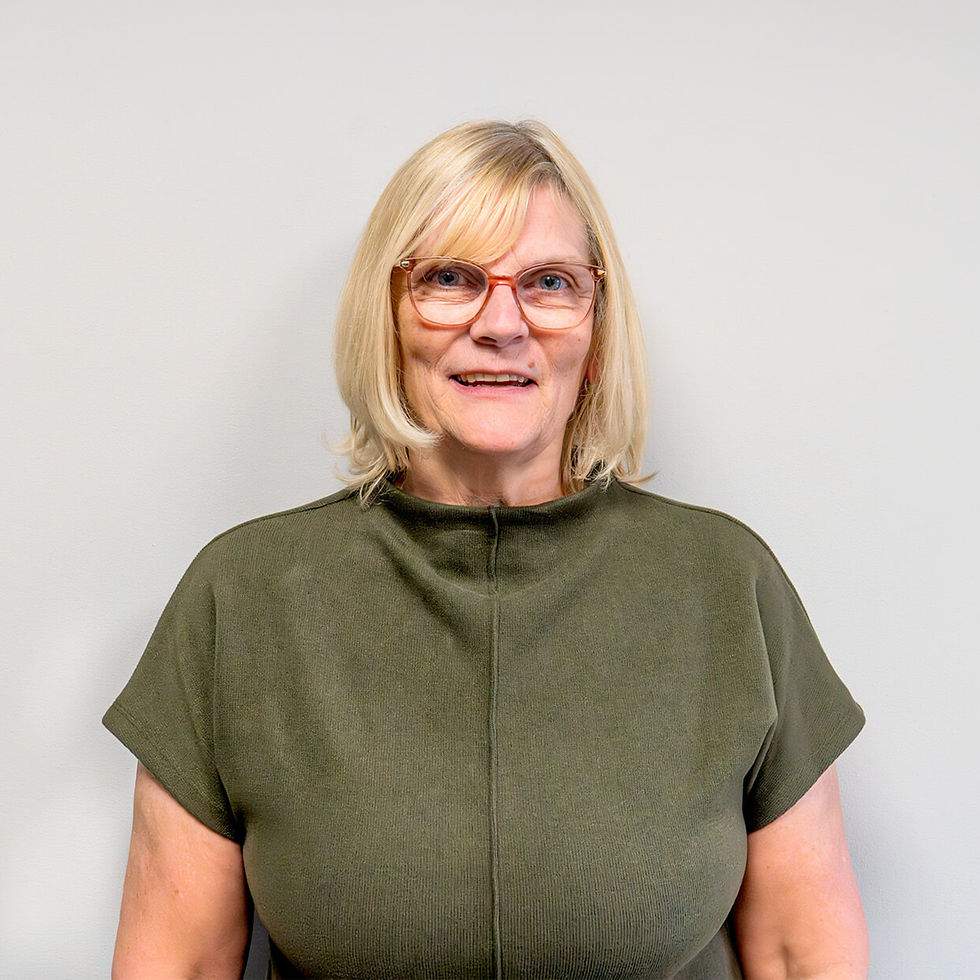 Portrait of a smiling blonde woman in glasses and olive top against a plain light wall.