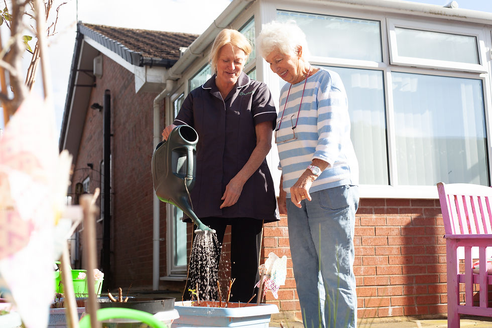 Two women gardening on a patio. One waters a potted plant with a watering can, both smiling. Sunlit brick house in the background.