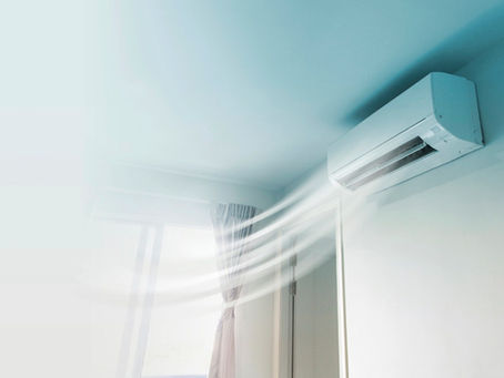 Indoor wall mounted air conditioner blowing cool air into a bright room with a window and curtain visible.