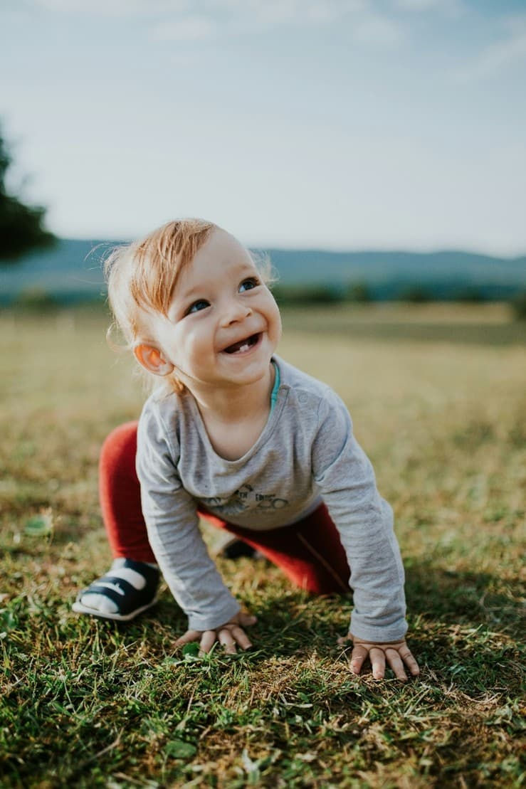A boy is crouching on the grass field, smiling with his small bottom teeth showing.
