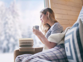 Woman sitting by a window with a hot drink and book, looking out at a snowy winter landscape.