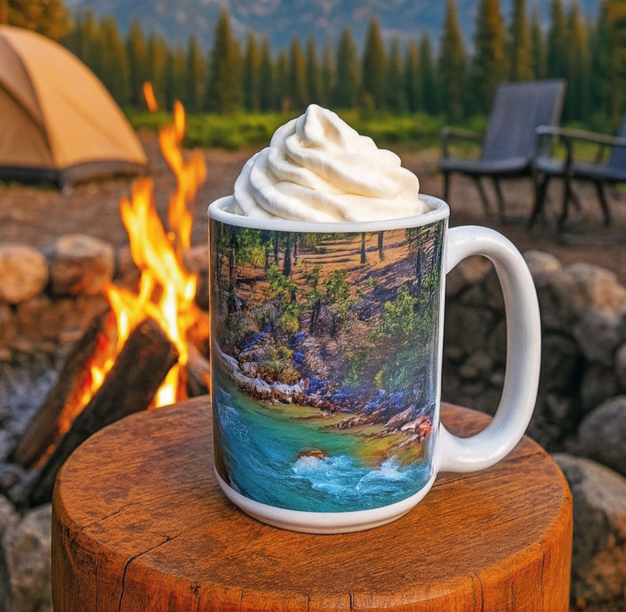 Thumbnail: 15 oz ceramic mug featuring a mountain river landscape photo, set on a wooden table beside a campfire with a tent, chairs, an