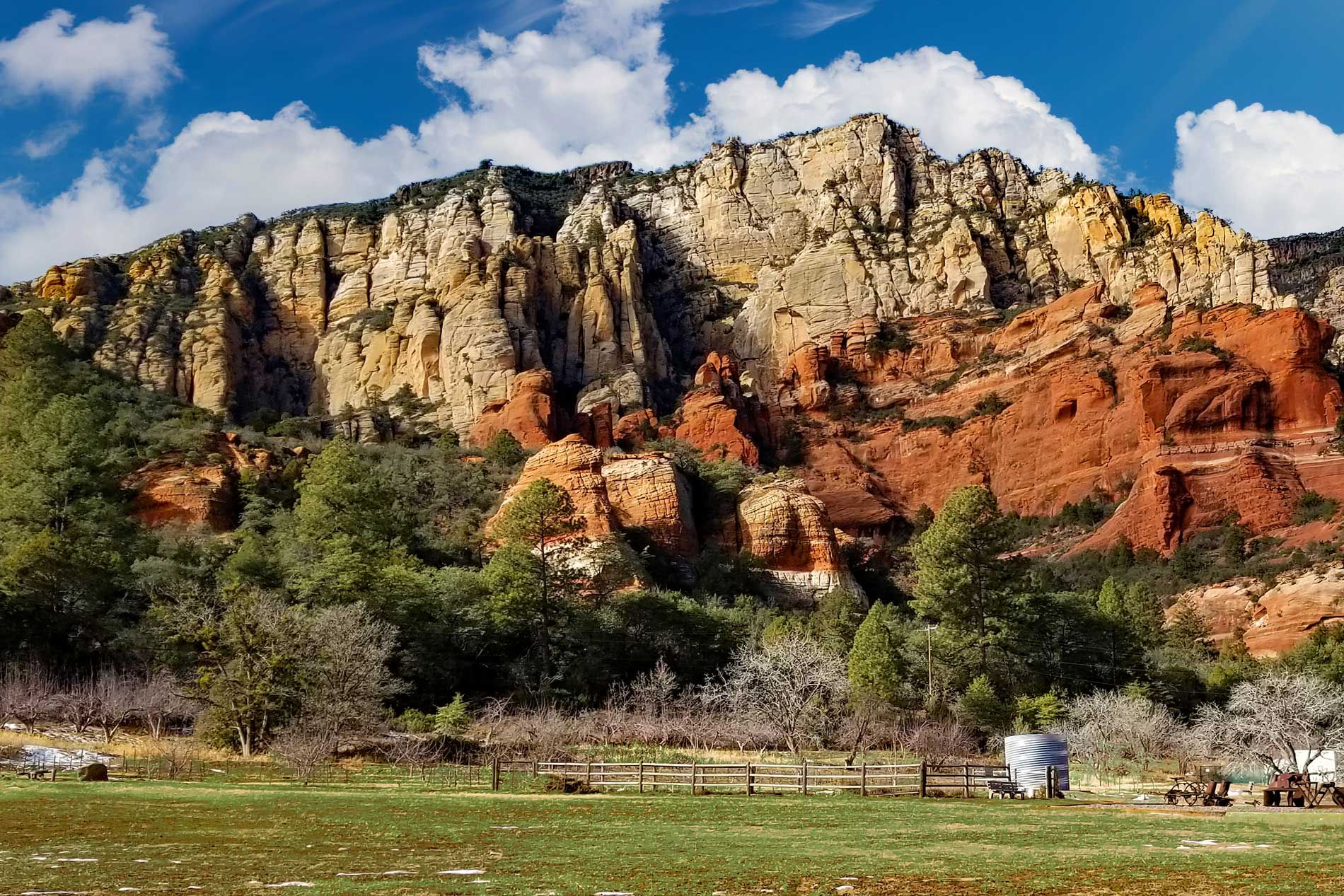 Layered red, orange, and cream sandstone cliffs rising above a green meadow with trees and fencing under a bright blue sky wi