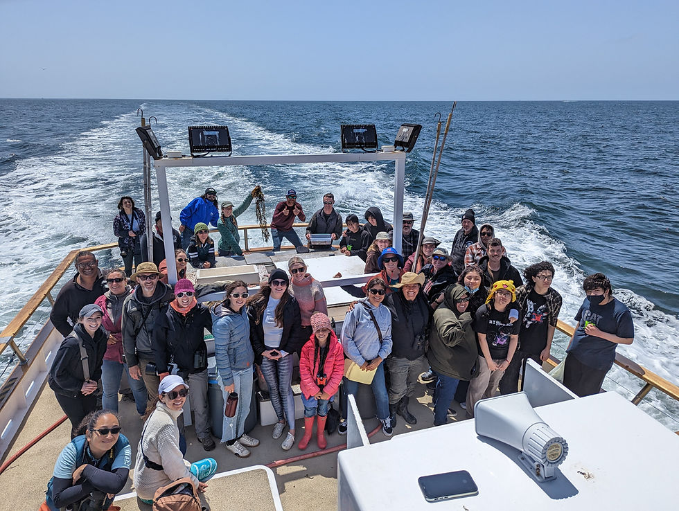 A group of about 30 people stand smiling on the back on a boat on a clear day. The boat's wake stretches to the horizon in the background.