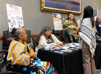 3 people behind an exhibitor booth are listening to a person standing in front of the booth. There are flyers and tripies with posters with information. 