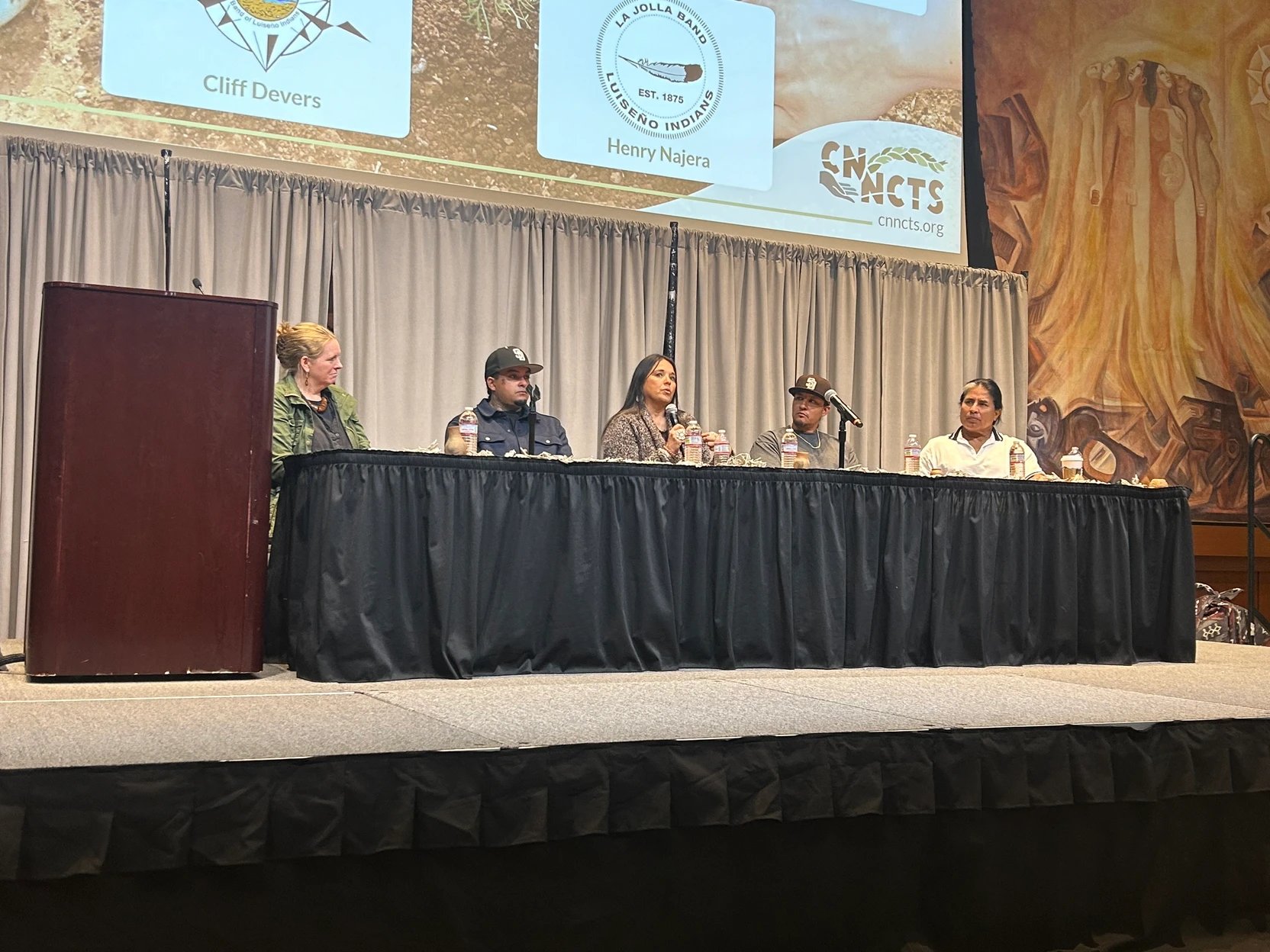 A panel of five people sit behind a table on a stage.
