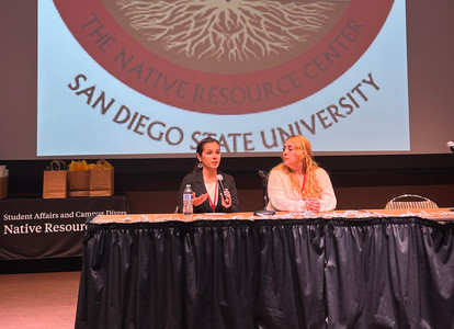 Two panelists are seated at a table with a black curtain, holding a microphone and speaking to the audience