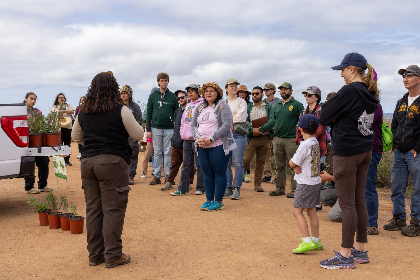 A person stands outside in front of a crowd talking. There are plants ready to be planted next to them.