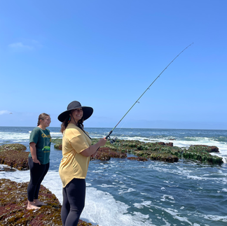 Two people stand at the coast. One holds a fishing rod in the water.