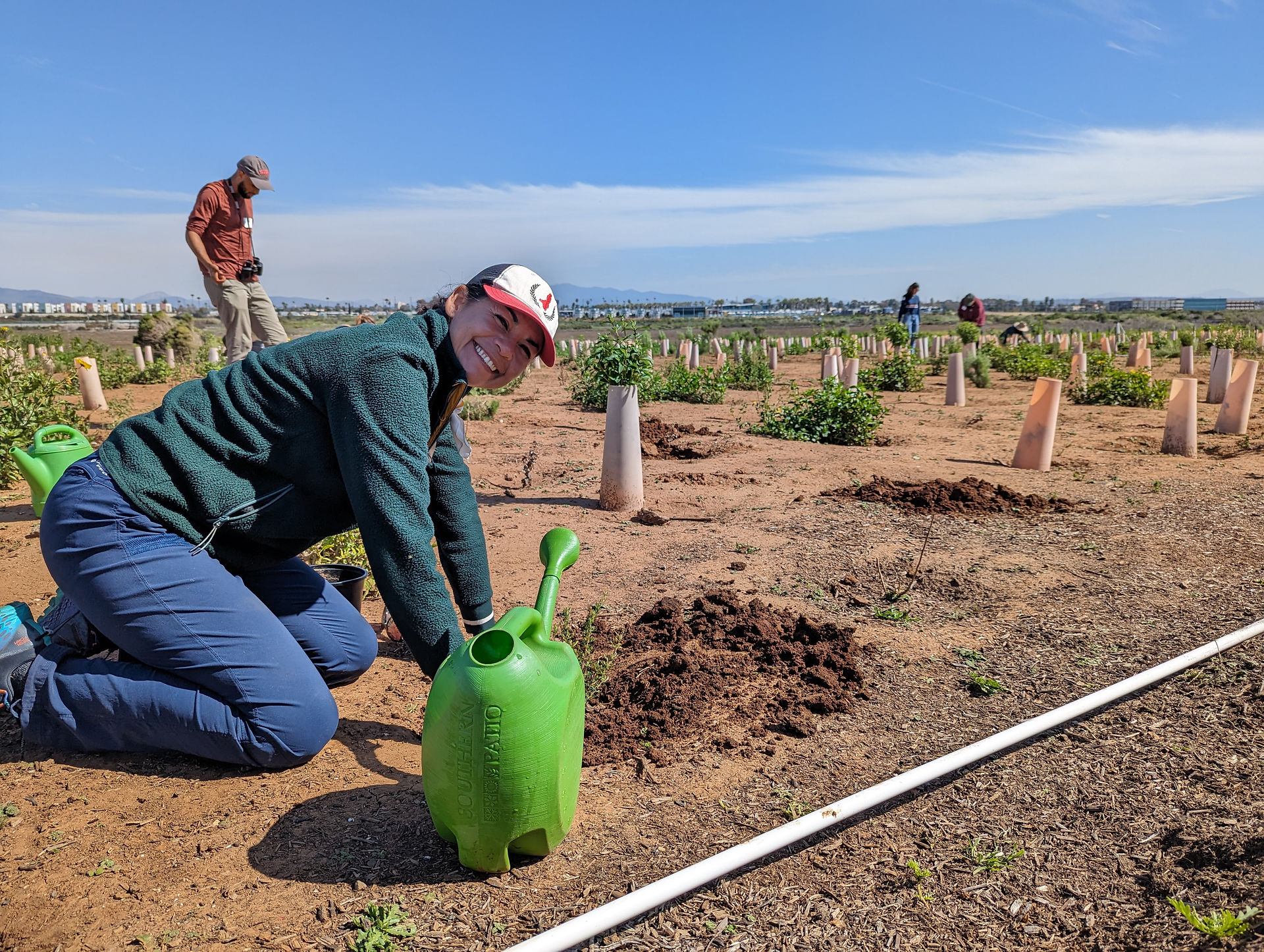 A participant smiles at the camera next to a watering can, while crouching and placing two hands in the soil