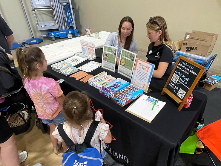 Two people sit behind the Climate Science Alliance booth, which includes a table with resources, art supplies, Clif bars, and information and a table set up with paper and pencils. Two children approach the table.