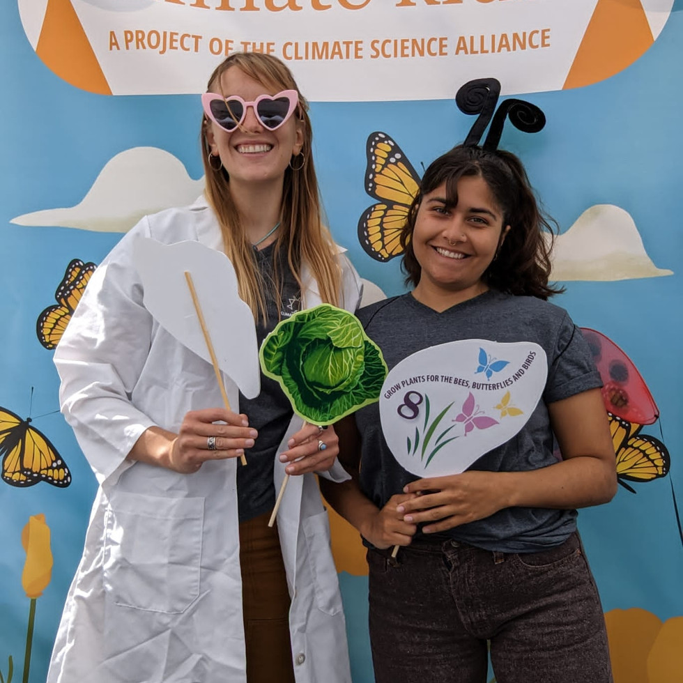 Two people smile in front of the Climate Kids photo booth, a blue backdrop with white clouds and yellow monarch butterflies and the Climate Kids logo. One person wears pink heart sunglasses and holds a lettuce prop. The other wears butterfly antenas and holds a butterfly prop.