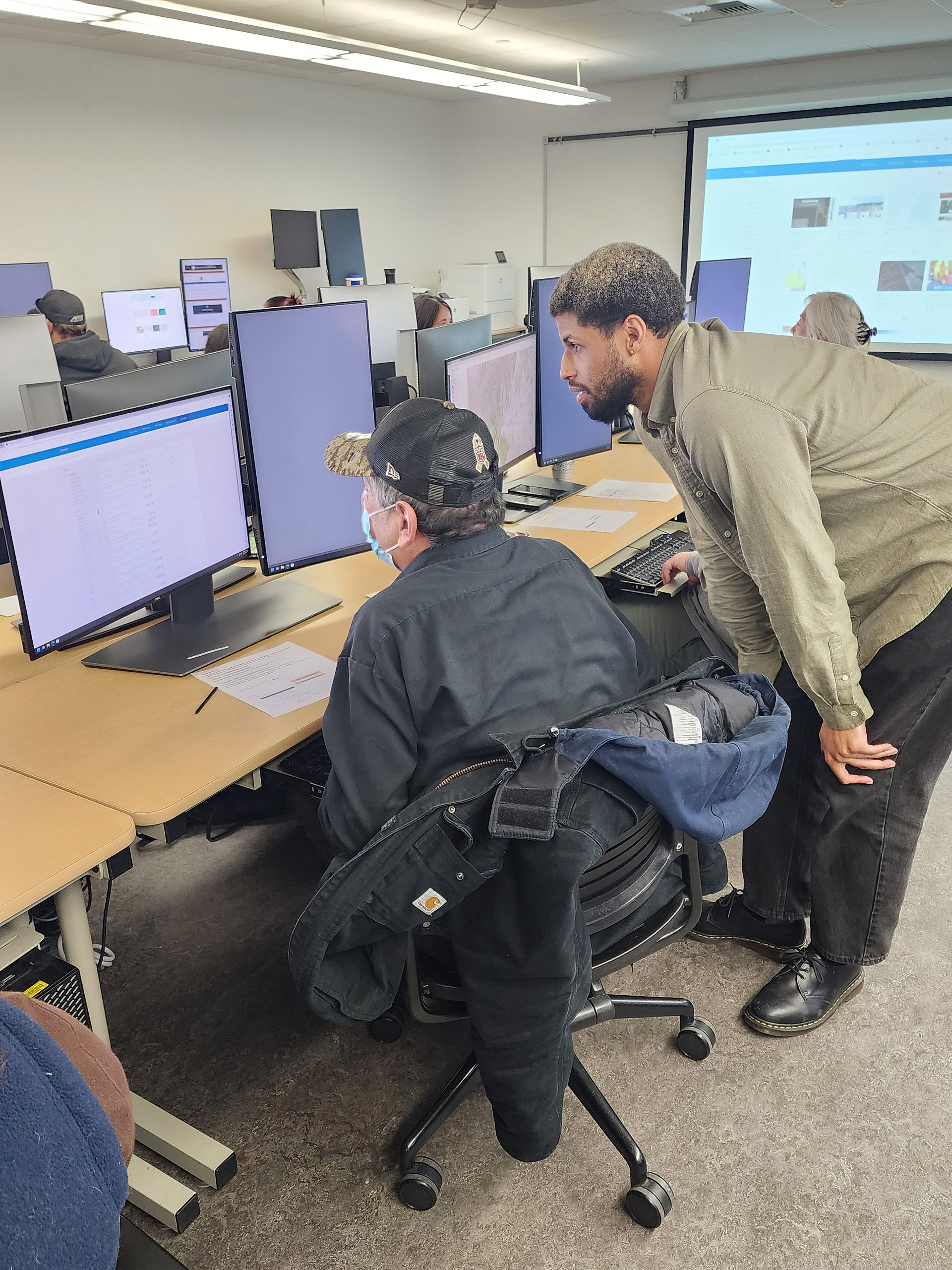 A person sits at a computer using ArcGIS software while another person stands beside them to assist.