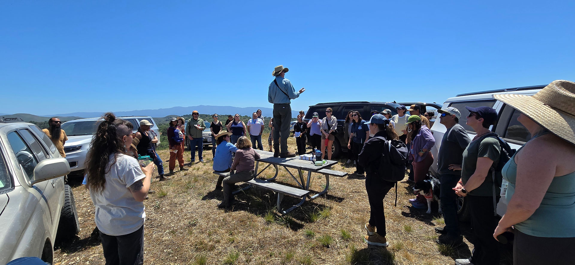A person stands on a picnic table talking to participants who are gathered around