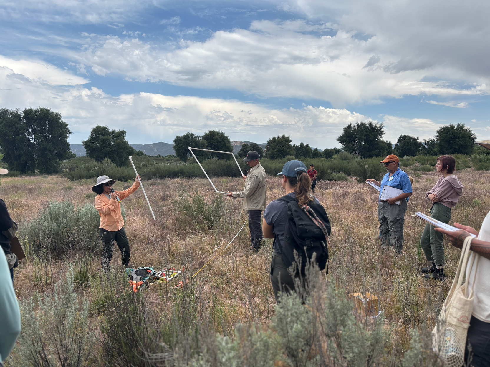 A group of people stand outside amid tall grass. Two people demonstrate with a square construction.