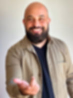Rapaz de barba, sem cabelo, de pele clara e cabelos castanhos escuros, vestindo uma camiseta azul escura por baixo de uma camisa de jeans aberta bege, atrás um fundo bege, ele sorri e estende a mão direita em direção a câmera