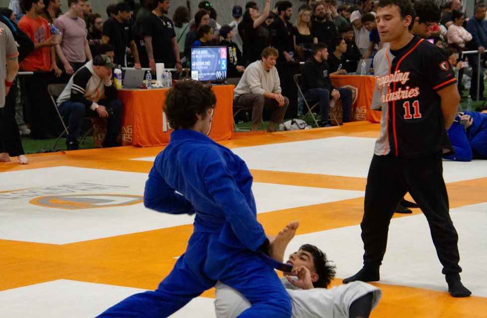 Blue-clad Boston jiu jitsugrappler pins a white-clad opponent on an orange mat; referee watches, crowd behind, with Grappling Industries jersey.