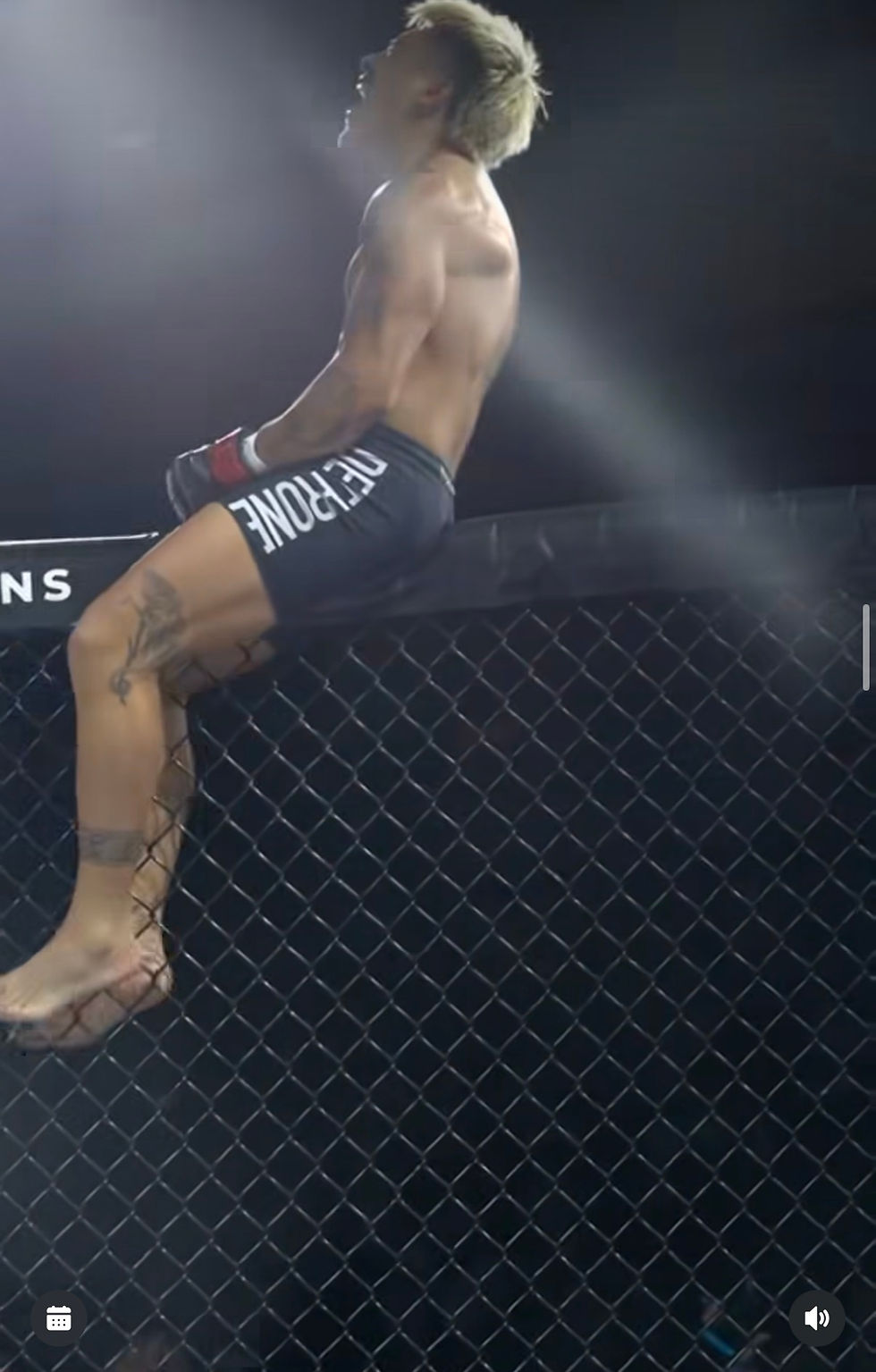 Shirtless Boston MMA fighter with bleached hair sits on the cage fence under bright lights, shorts reading FERONE.