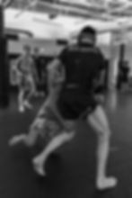Black-and-white gym scene of two men grappling on martial arts Boston = mats, with BACK TO BASICS on one shirt and others watching in background.