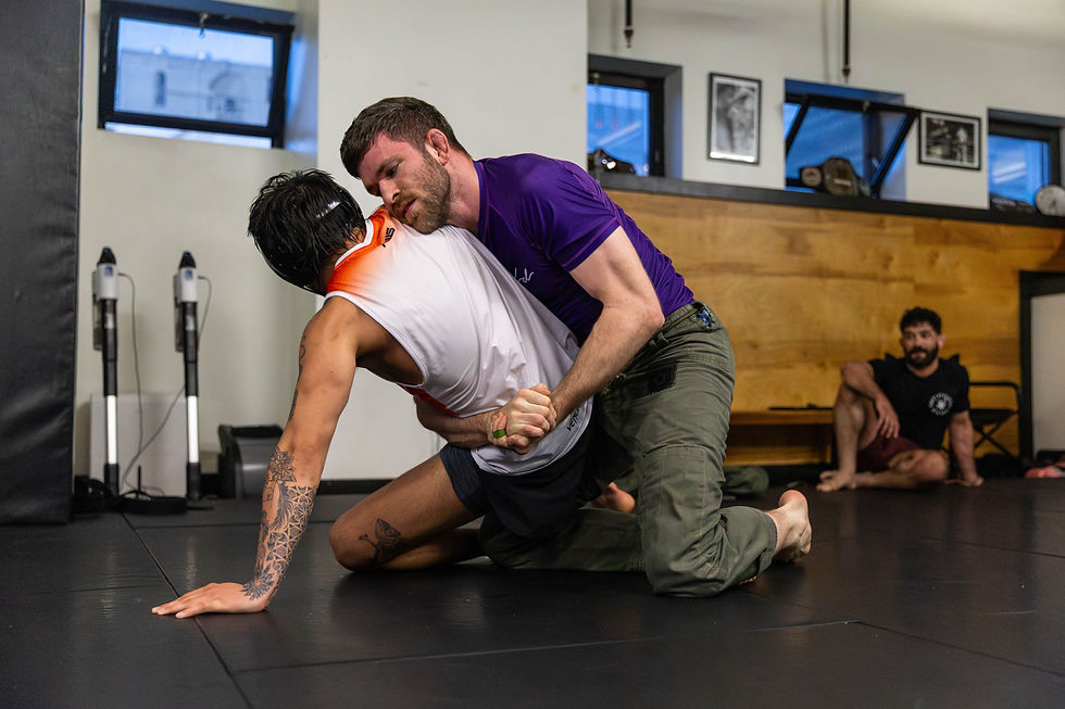 Two men grapple on a Boston jiu jitsu gym mat, one in a white shirt, the other in purple. A seated man observes. Gym equipment and framed photos adorn the walls.
