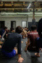 A group of Boston Jiu Jitsu people in athletic wear sit on a gym mat, attentively listening to an instructor. Wood beam ceiling, "BACK 2 BASICS" text visible.