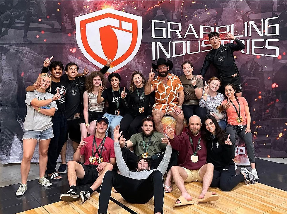 Group of Boston jiu jitsu practitioners pose and cheer in front of a Grappling Industries banner, smiling and making victory gestures.