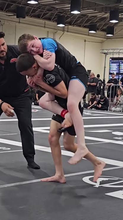 Two boys grapple on a mat in a gym, one carrying the other on his back while a referee watches. ADCC is greatly loved by Boston Jiu Jitsu practitioners.