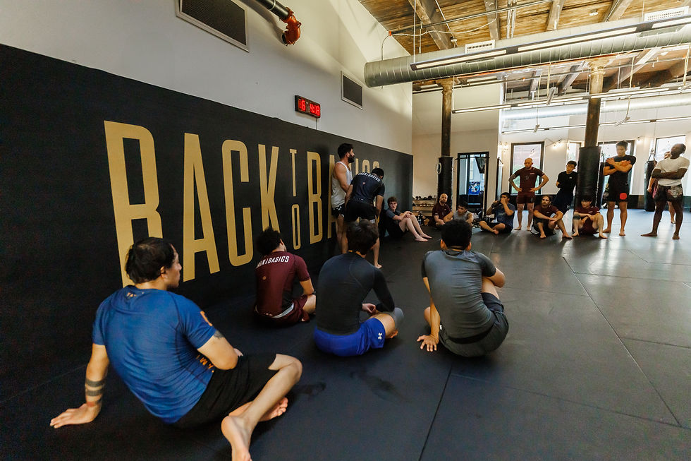 MMA class gathering on  Boston jiu jitsu black mats in an industrial gym, students seated and listening under a large BACKBASICS wall sign.