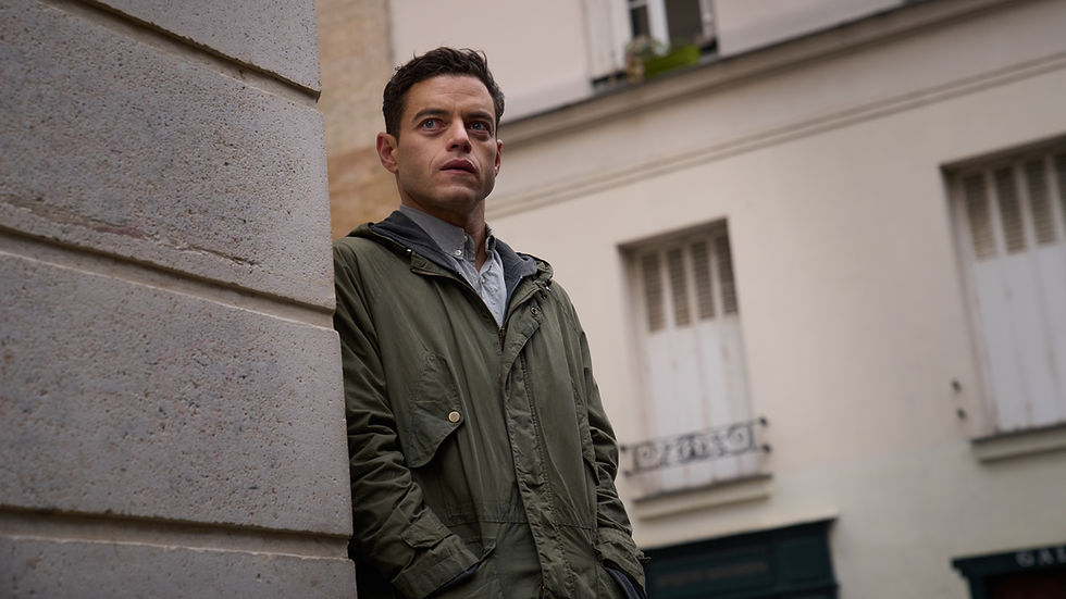 A man dressed in a green coat leans against a stone wall and appears to be watching something or someone.