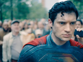 Hero in blue and red suit walks through a crowded street, people taking photos. Focused expression, blurred green trees and people behind.
