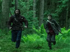 Two people run through a dense, green forest carrying bows. They wear dark clothing and appear determined and focused amidst the tall trees.