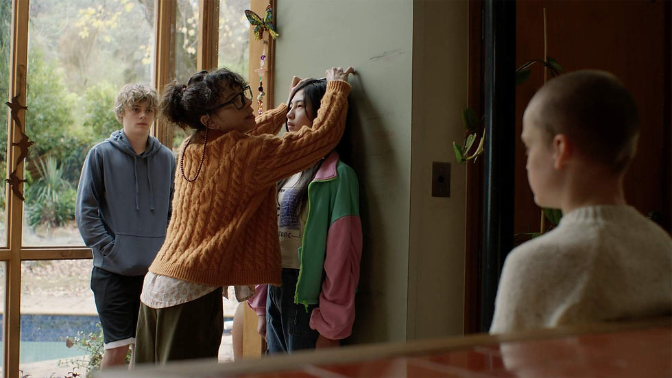 A woman measures a girl's height against a wall in a cozy room. A boy in a hoodie watches, with a leafy garden visible outside.