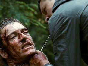 Two men in a tense scene in a forest, one covered in blood, held by a chain. The mood is intense and confronting. Dense greenery behind.