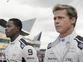 Two men in racing suits stand focused at a racetrack. The cloudy sky and modern architecture form the backdrop. Logos on suits are visible.