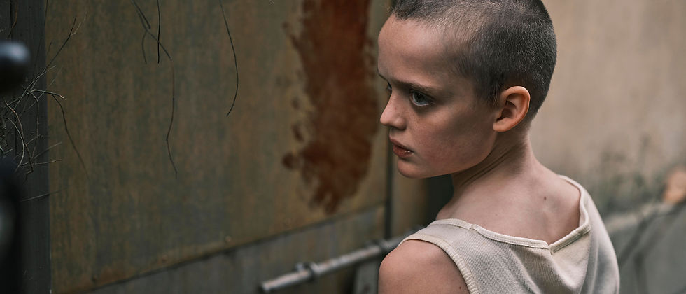 A child with a shaved head in a white tank top stands by a rusty wall, looking back with a serious expression. The mood is tense.