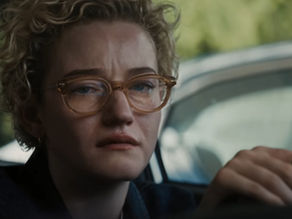 Woman with glasses looks distressed, sitting in a car. Blurred green background through the window suggests an outdoor setting.