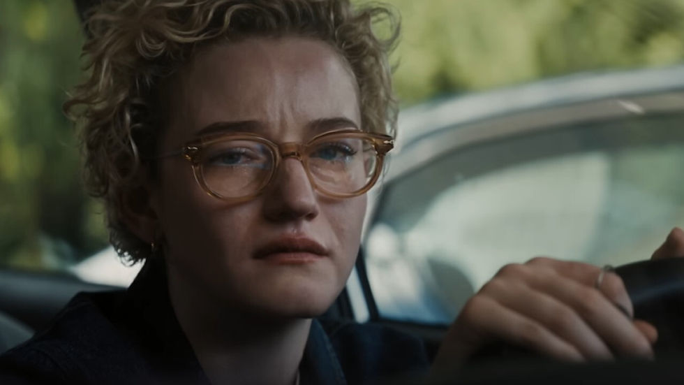 Woman with glasses looks distressed, sitting in a car. Blurred green background through the window suggests an outdoor setting.