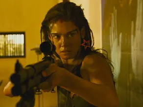 A woman aims a rifle intensely in a dimly lit room. She has a focused expression, dark hair, and wears an pink star earring.